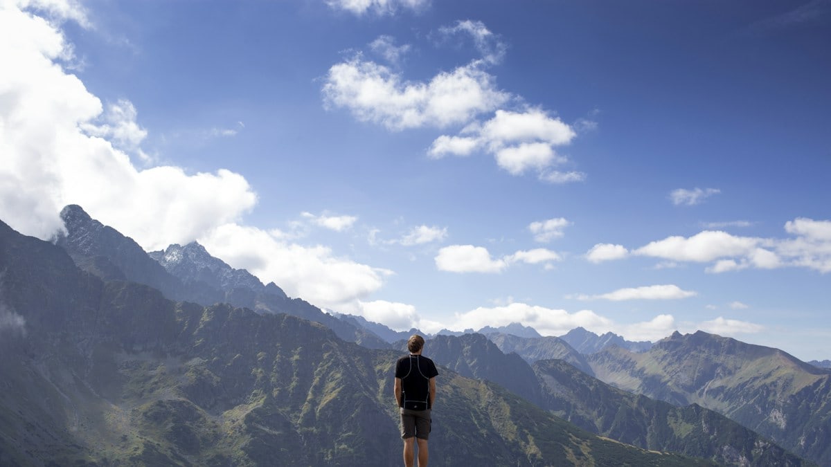 man wearing black shirt and gray shorts on mountain hill beside mountains under white and blue cloudy skies